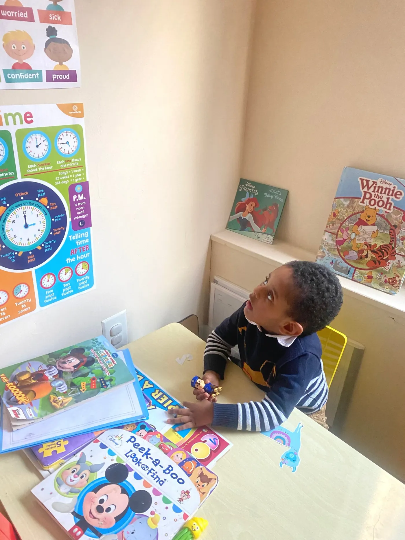 A young boy at a child-sized table in the daycare's learning corner, surrounded by picture books and puzzles, looking up at a colorful poster about telling the time.