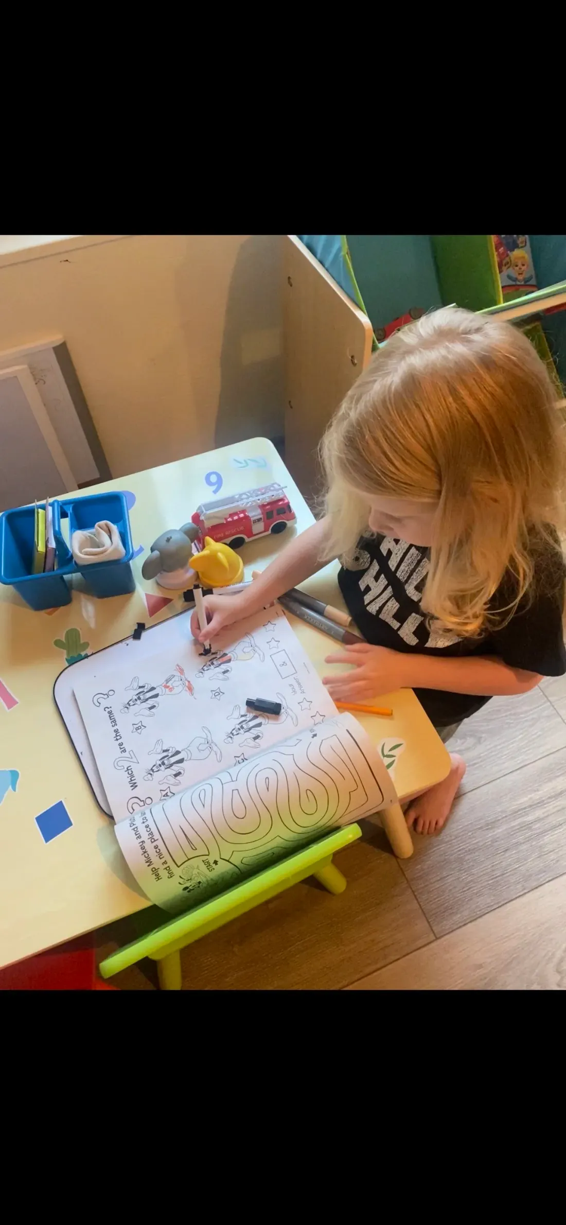 A young girl at a play table working through a printed activity book, holding a marker over a matching puzzle that asks which two pictures are the same.